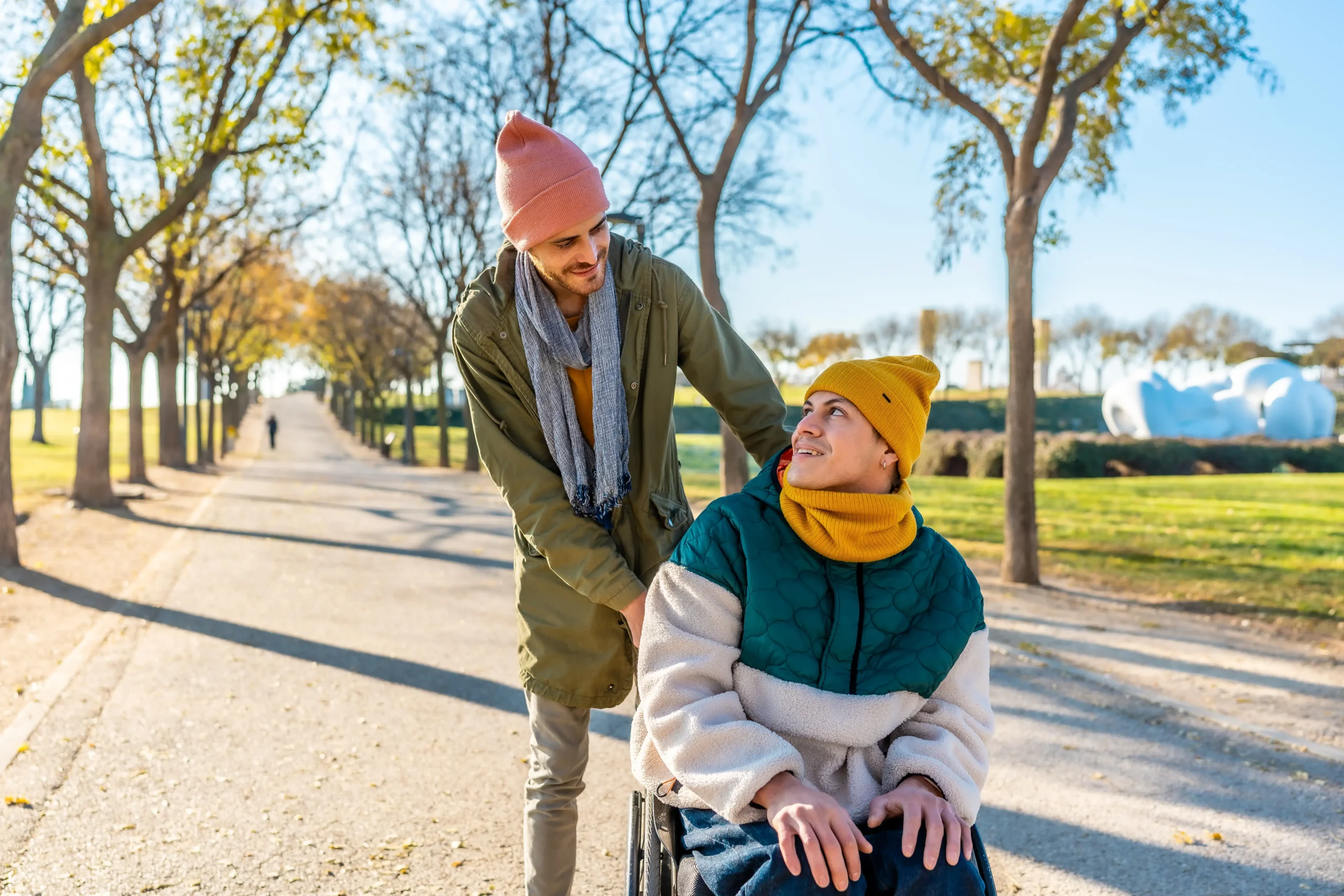 A sunny outdoor scene in a park with autumn trees. One young man is sitting in a wheelchair, wearing a colorful puffer jacket and a yellow beanie, looking up and smiling at his companion. The other man stands behind him, leaning down with a smile, wearing an olive parka, a gray scarf, and a pink beanie.