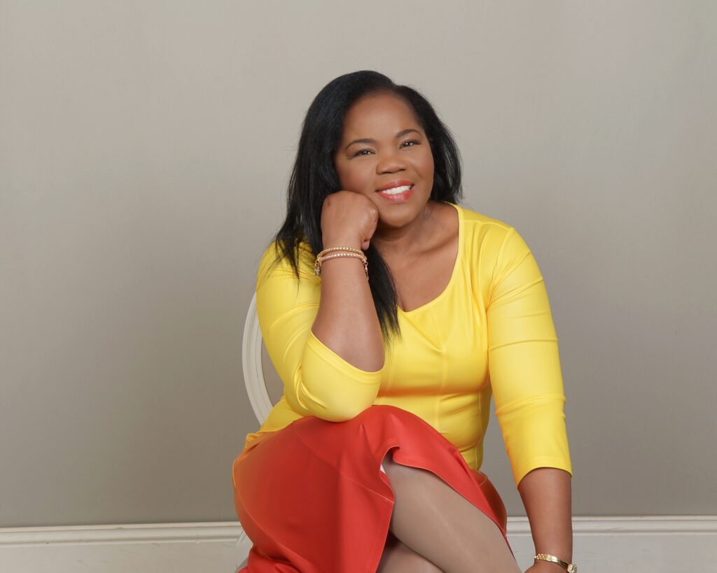 A professional portrait of a woman with long dark hair, wearing a vibrant yellow top and a red skirt. She is sitting on a white chair against a neutral grey wall, leaning forward with her chin resting on her hand and smiling warmly at the camera.