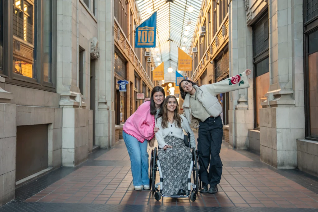 Three young women are smiling and posing together in a brightly lit, historic indoor shopping arcade with a glass ceiling. One woman is seated in a manual wheelchair wearing a long patterned skirt and a white cardigan. She is flanked by two friends; one in a pink sweater leaning in close, and another in a cream-colored puffer jacket with her arm outstretched in a joyful gesture. Colorful banners hang from the balconies above them in the background.