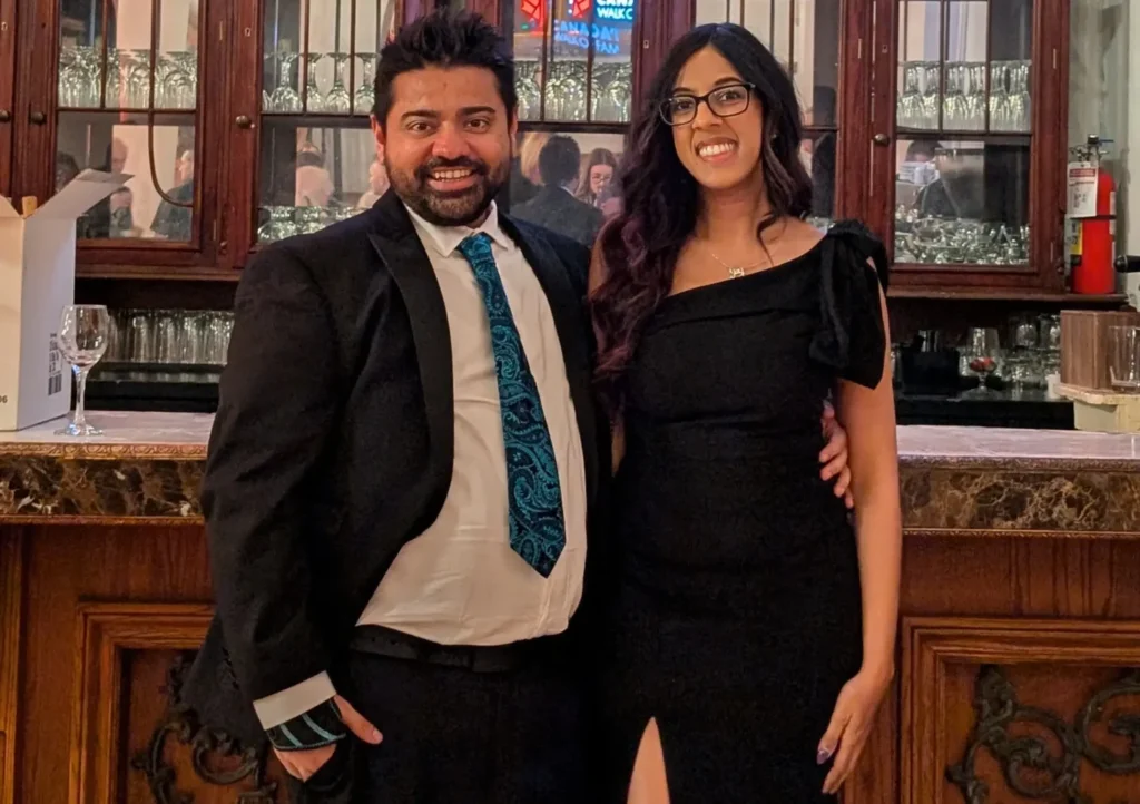A formal photo of a couple posing in front of a dark wood bar with ornate glass cabinets. Above them is a glowing neon sign that reads "Canada's Walk of Fame" with a maple leaf logo. The man wears a black suit with a patterned teal tie. The woman wears a floor-length, one-shoulder black gown with a high leg slit and glasses. Both are smiling warmly at the camera.