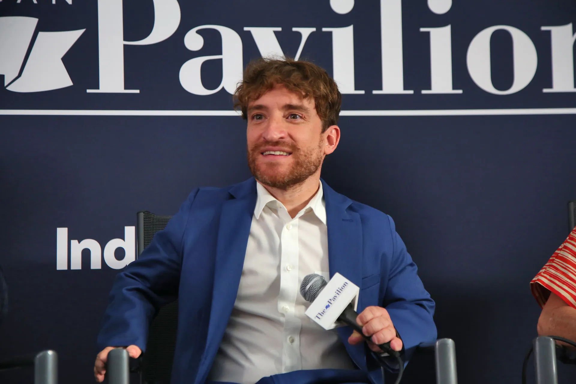 A man with wavy light-brown hair and a groomed beard is seated and speaking into a microphone during a panel or interview. He is wearing a sharp blue suit over a crisp white button-down shirt. Behind him is a dark blue backdrop with the word "Pavilion" in large white serif lettering. He has an engaged, slightly smiling expression as he looks off-camera.