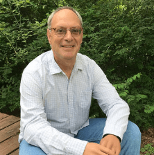 A portrait of a man with short grey hair and glasses, smiling while seated outdoors. He is wearing a light-colored, thin-checked button-down shirt and blue jeans. He is positioned in front of a dense backdrop of lush green bushes and trees, with a wooden deck visible to the left.