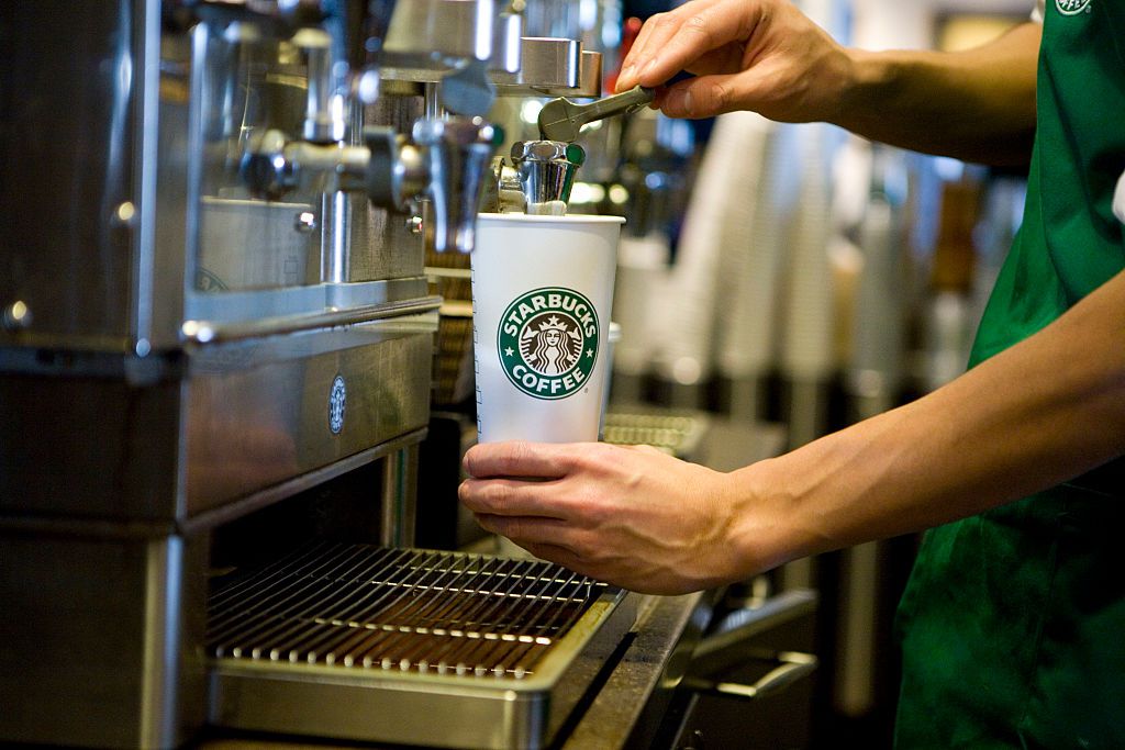 A close-up, action shot of a barista's hands preparing a drink. One hand holds a white Starbucks paper cup with the classic green siren logo under a metal dispenser, while the other hand operates a lever. The background is slightly blurred, showing the industrial textures of the espresso machine and a stack of cups, emphasizing the focus on the pour.