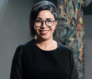 A head-and-shoulders portrait of a woman with short, dark hair and glasses, wearing a black top. She is smiling warmly at the camera against a soft-focus background that includes a colorful, abstract art piece.