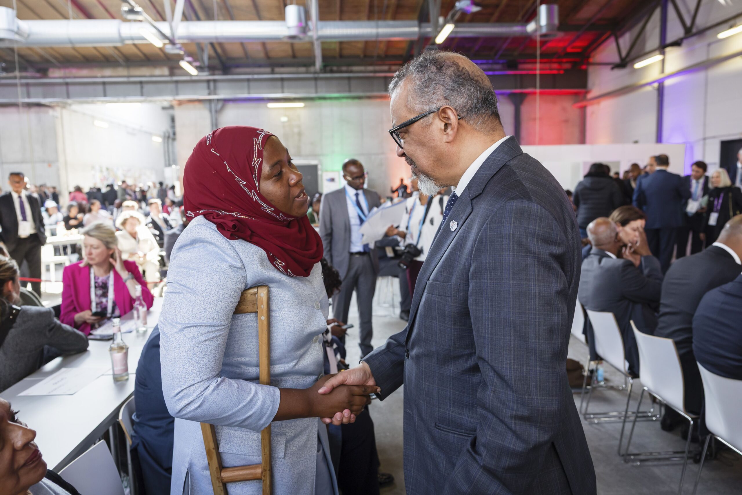 A candid shot of two people shaking hands in a large, busy indoor event space. On the left, a woman wearing a red hijab and a light blue long-sleeved dress uses a wooden crutch for support while shaking hands. On the right, a man in a grey checkered suit and glasses smiles warmly at her. The background is filled with other attendees sitting at long tables under bright, industrial-style ceilings.