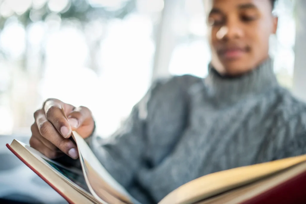 A close-up, shallow-focus shot of a man's hand turning a page of a red hardcover book. The man, partially blurred in the background, wears a grey cable-knit turtleneck sweater and has a soft, focused expression.