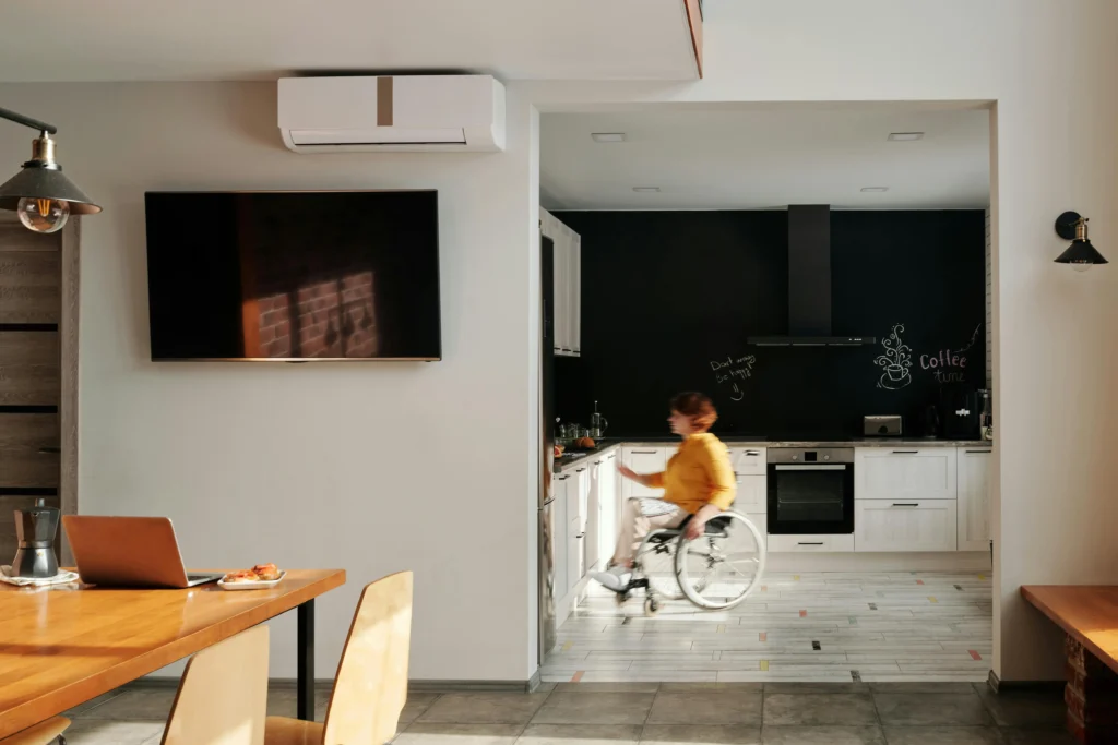 A person in a wheelchair is navigating a modern, open-concept kitchen. They are wearing a yellow shirt and moving toward a white kitchen counter. The kitchen features white cabinetry, a black backsplash with chalkboard art, and a stainless steel oven. In the foreground, a wooden dining table holds a laptop and a small plate of food, while a large flat-screen TV and an air conditioning unit are mounted on the adjacent white wall.