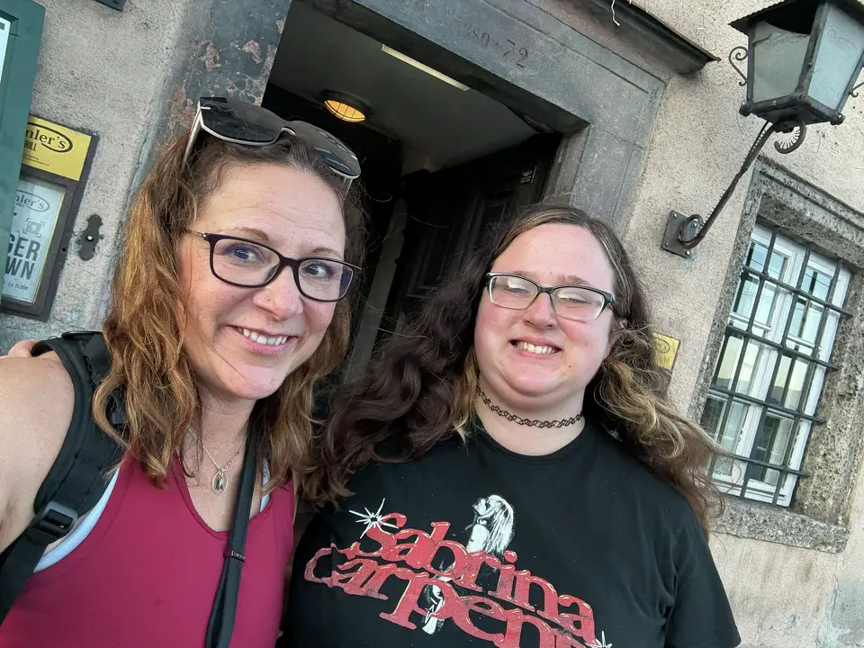 A casual outdoor selfie of two women smiling at the camera. The woman on the left has reddish-brown wavy hair, wears glasses, and a burgundy tank top with a backpack strap visible. The woman on the right has long, dark curly hair, wears glasses, a black "Sabrina Carpenter" graphic t-shirt, and a black lace choker. They are standing in front of a stone building with a vintage-style lantern and a window with a metal grate.