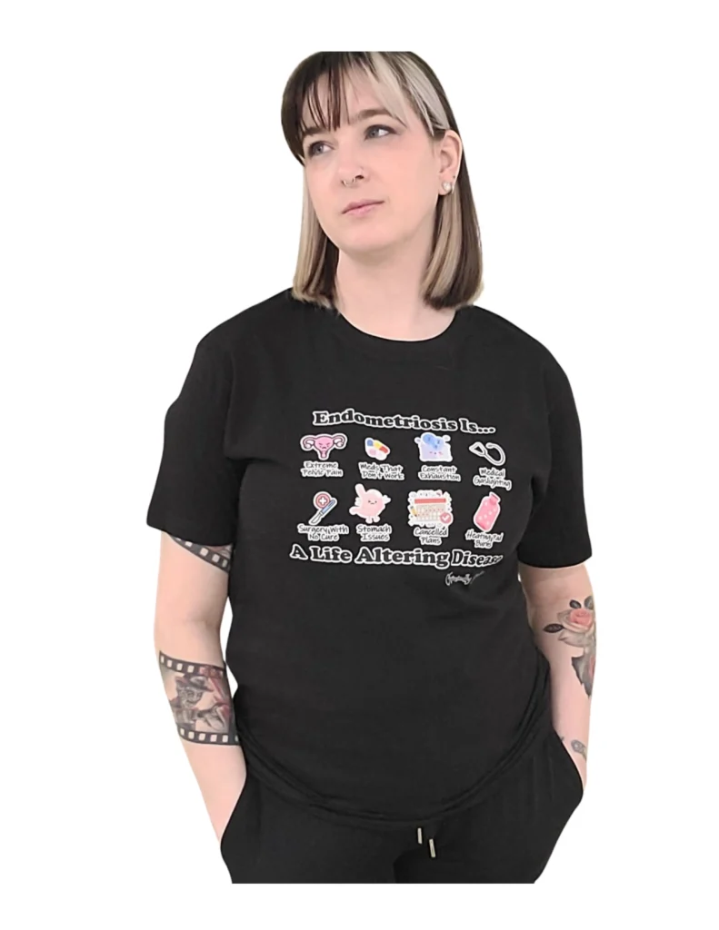 A woman with blonde and brown hair and arm tattoos wearing a black t-shirt. The shirt features a grid of eight cartoon icons illustrating endometriosis symptoms, including "Extreme Pelvic Pain," "Constant Exhaustion," and "Medical Gaslighting." Large text at the top and bottom reads, "Endometriosis Is... A Life Altering Disease."