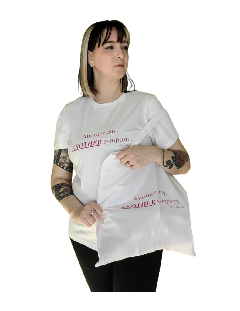 A woman with tattoos on both arms wearing a white t-shirt and holding a matching white tote bag. Both the shirt and the bag feature the phrase "Another day, ANOTHER symptom." printed in a simple serif font, with the word "ANOTHER" emphasized in a bold, dark red underline.