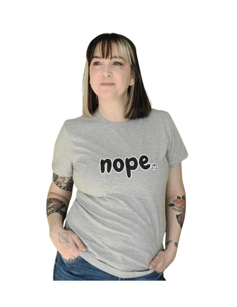 A woman with a nose ring and tattooed arms posing in a grey t-shirt and blue jeans. The shirt features the word "nope." in a bold, rounded black font with a thick white outline. A small penguin logo is positioned just to the right of the text.