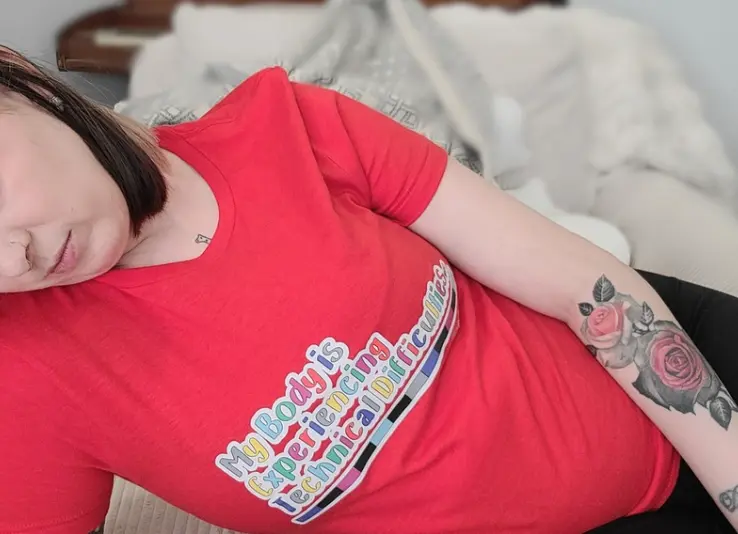 A close-up shot of a person reclining, wearing a vibrant red t-shirt. The shirt displays the text "My Body is Experiencing Technical Difficulties..." in a playful, colorful font outlined in white, set against a backdrop of a colorful horizontal test pattern bar. A rose tattoo is visible on the person’s forearm.