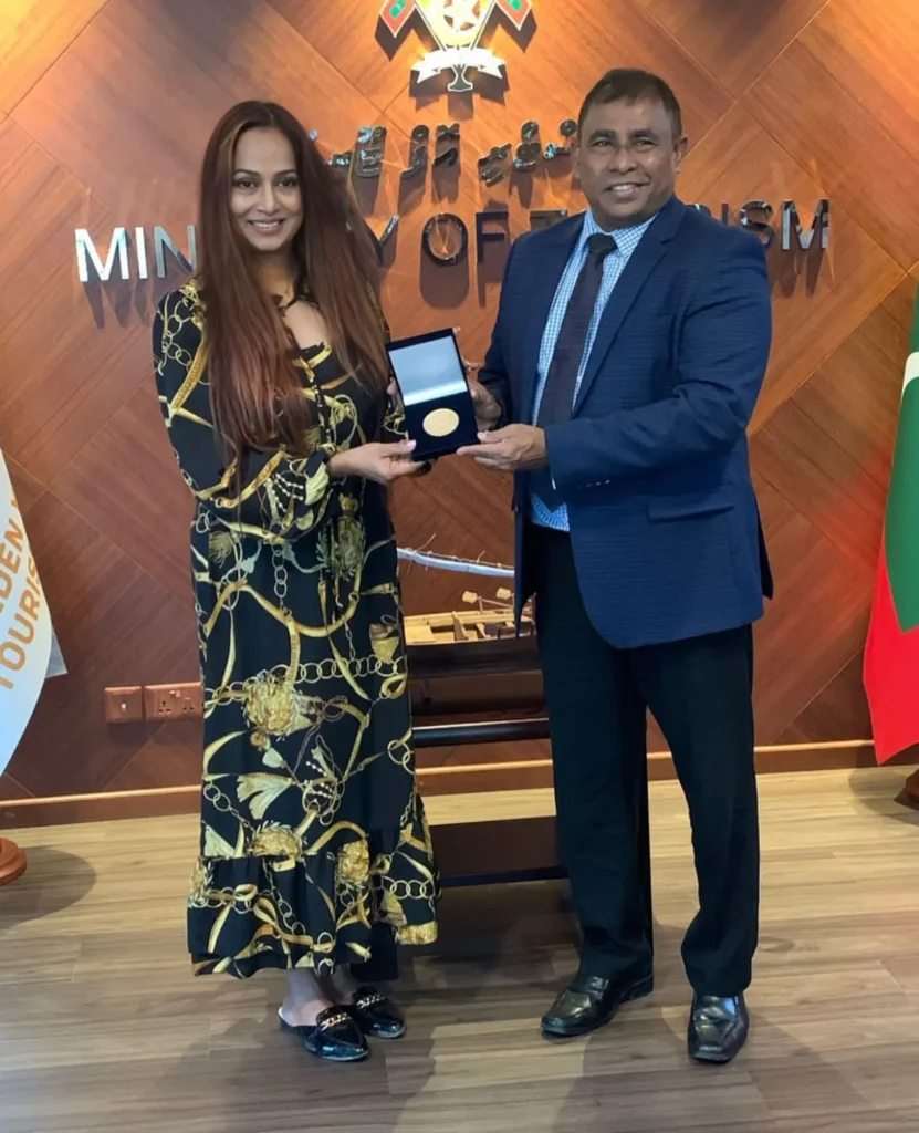 A woman in a black and gold patterned dress receiving a gold medal in a display case from a man in a blue suit at the Ministry of Tourism.