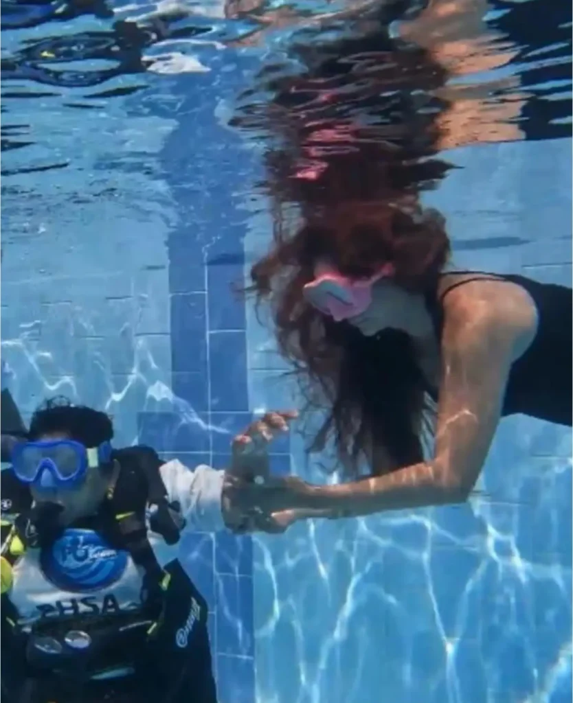 A female diving instructor with long hair and a pink mask assisting a scuba diver wearing a HSA (Handicapped Scuba Association) vest underwater in a pool.