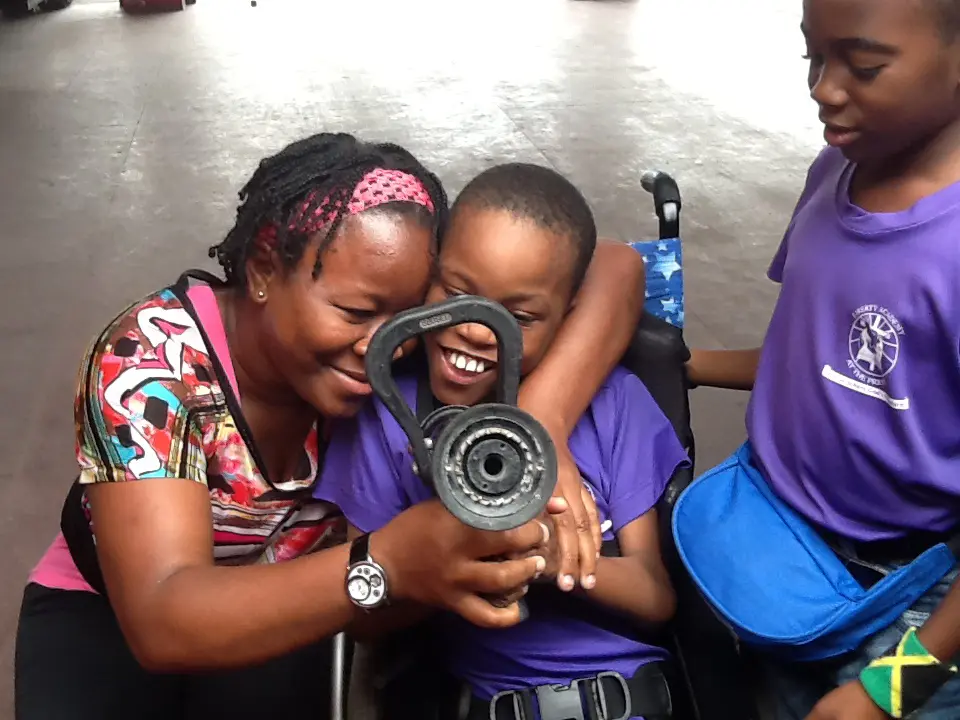 A woman with a pink patterned headband smiles warmly while leaning in close to a laughing young boy in a wheelchair. A third person in a purple shirt stands beside them, and the group appears to be interacting with a piece of equipment in an indoor setting.