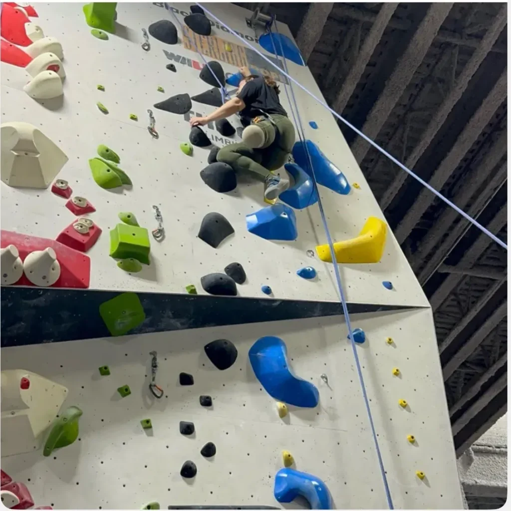 An adaptive athlete using a prosthetic leg climbs an indoor rock wall featuring various colorful holds and safety ropes.