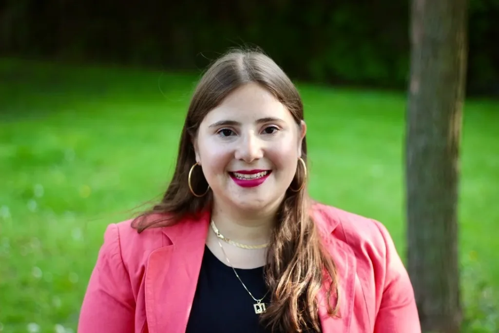 A smiling woman with long brown hair, wearing a pink blazer over a black top and gold hoop earrings, posing in a lush green park.