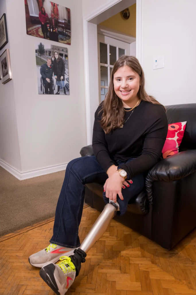 A young woman with a prosthetic leg sits comfortably in a black leather armchair in a brightly lit room with hardwood floors.