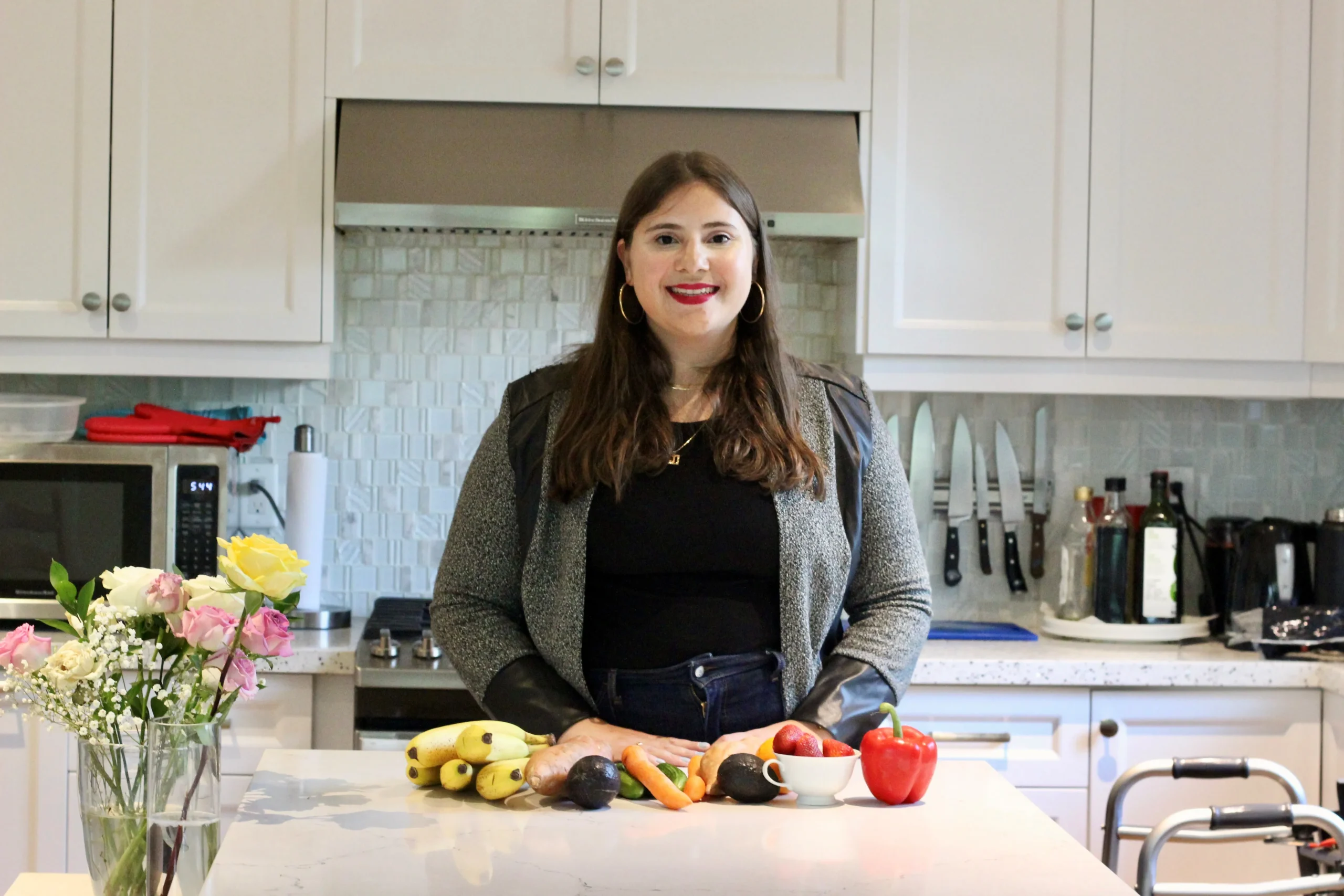 A woman stands behind a kitchen island filled with fresh produce including bananas, peppers, and avocados, in a modern kitchen with white cabinetry.