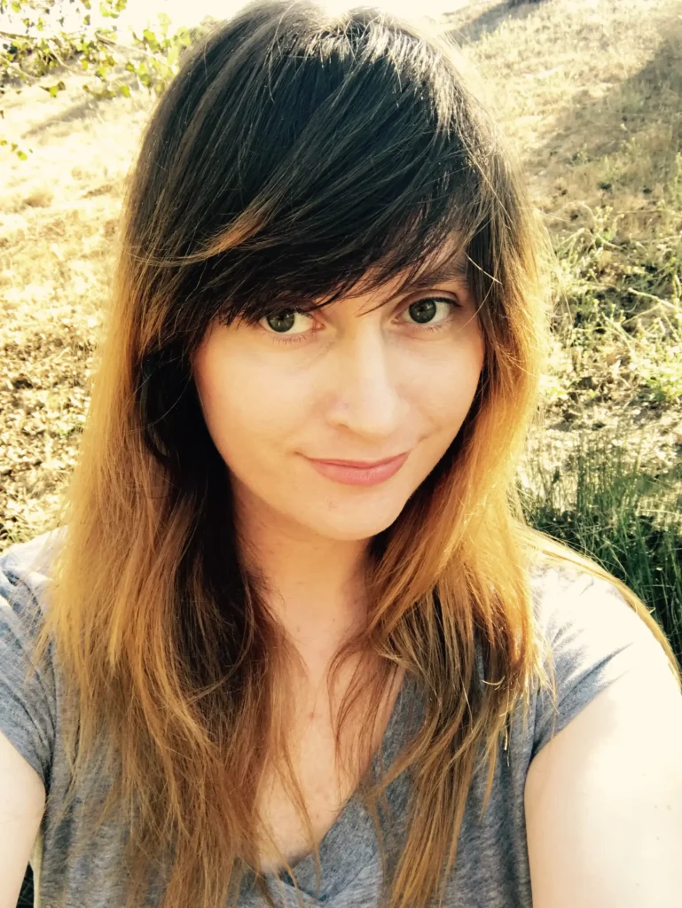 A portrait of Jessica Allen, a woman with long ombre hair—dark brunette at the roots transitioning to golden blonde at the ends—and a side-swept fringe. She is wearing a grey V-neck shirt and smiling softly in an outdoor setting with a sun-drenched, grassy hill in the background.
