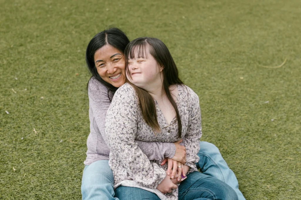 A happy mother and her teenage daughter with Down syndrome sharing a warm embrace while sitting together on a green lawn outdoors.