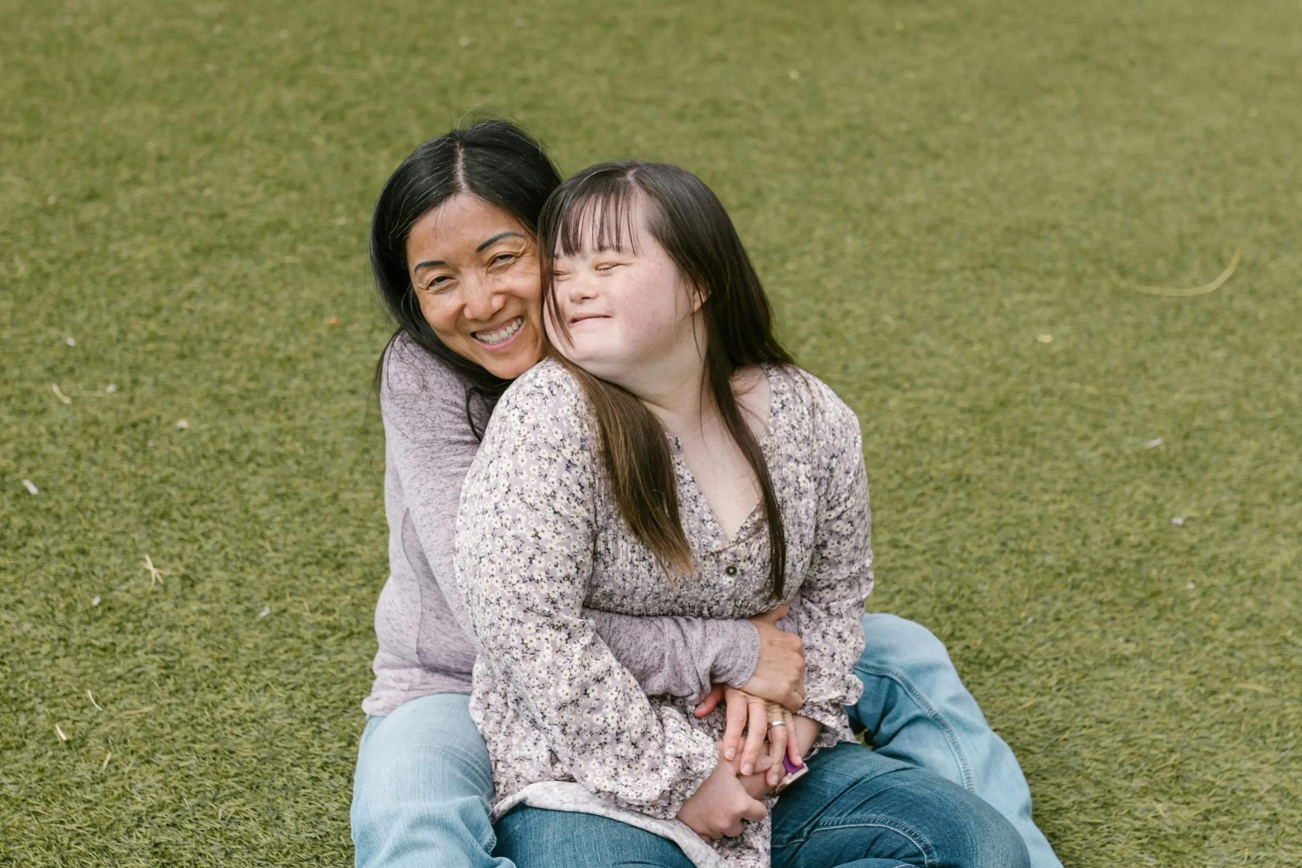A happy mother and her teenage daughter with Down syndrome sharing a warm embrace while sitting together on a green lawn outdoors.