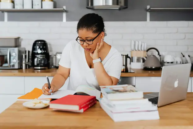 A focused woman wearing glasses and a white t-shirt sitting at a kitchen island, writing in a notebook surrounded by a laptop and stacks of textbooks.