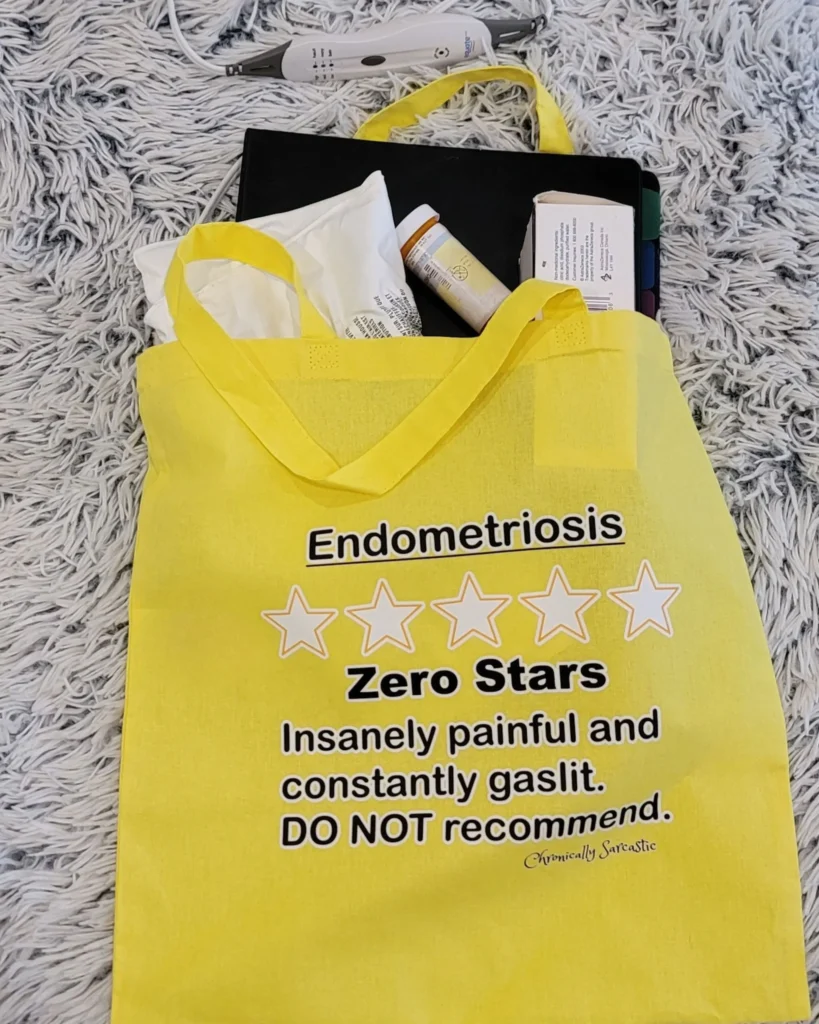 A yellow tote bag resting on a white shag rug, filled with medical supplies like pill bottles and a thermometer. The bag is printed with a "Zero Stars" review for Endometriosis, featuring five empty star outlines and the text: "Insanely painful and constantly gaslit. DO NOT recommend."