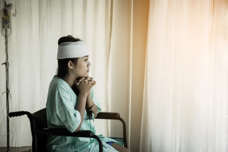 A young female patient in a light green hospital gown and a white head bandage sitting in a wheelchair, looking thoughtfully out of a bright window in a clinical setting.