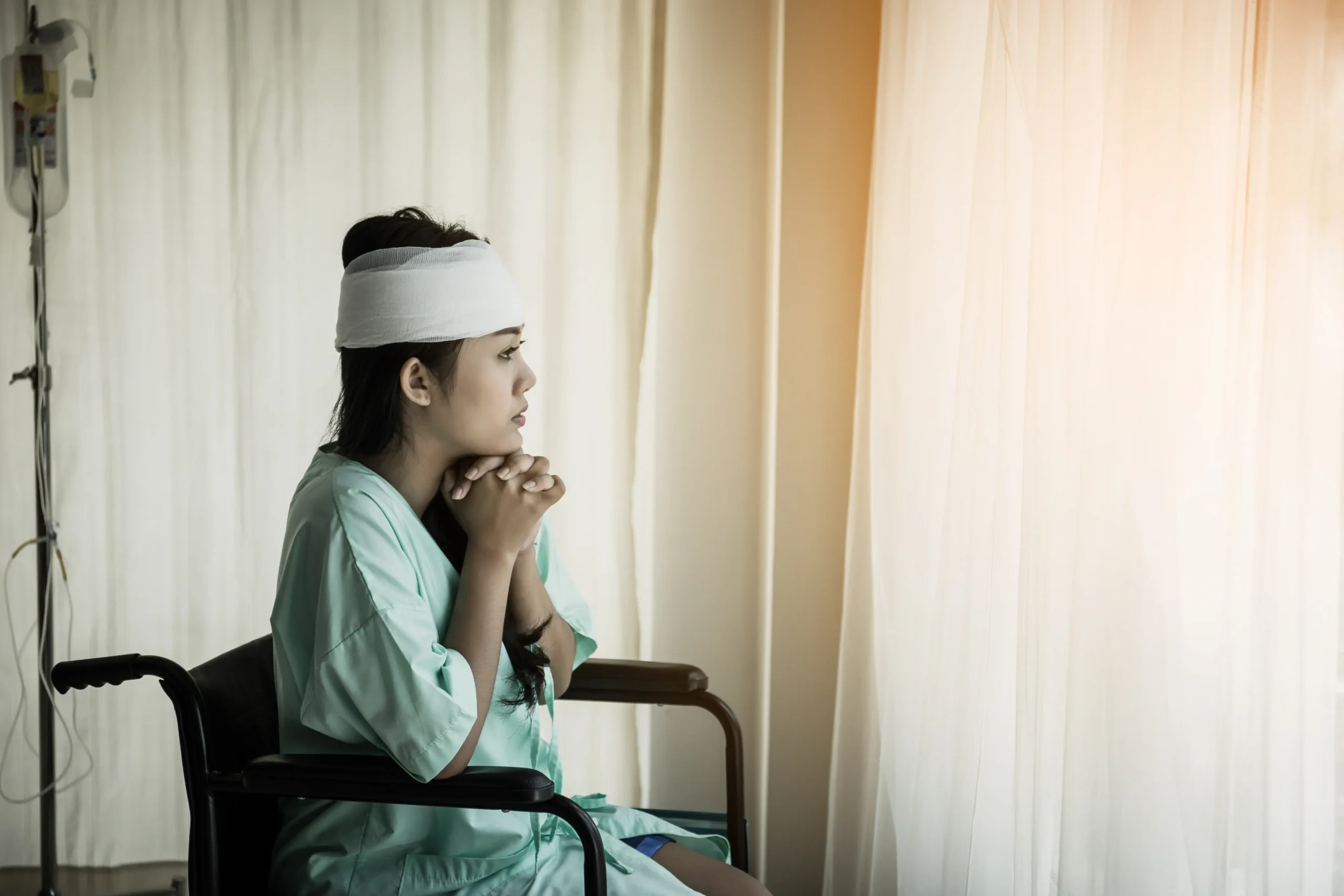 A young female patient in a light green hospital gown and a white head bandage sitting in a wheelchair, looking thoughtfully out of a bright window in a clinical setting.