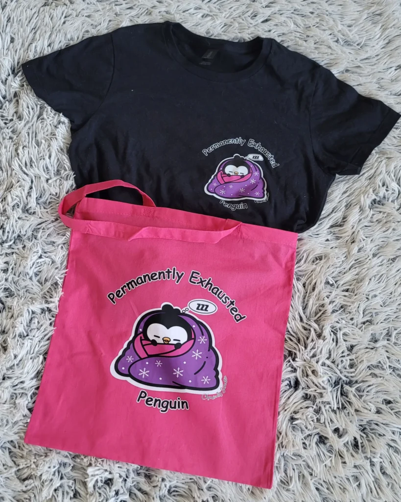 A flat-lay composition on a white shag rug featuring a black t-shirt and a bright pink tote bag. Both items are decorated with a graphic of a sleepy penguin wrapped in a purple blanket with snowflake patterns, surrounded by the words "Permanently Exhausted Penguin" and "ZZZ" in a thought bubble.