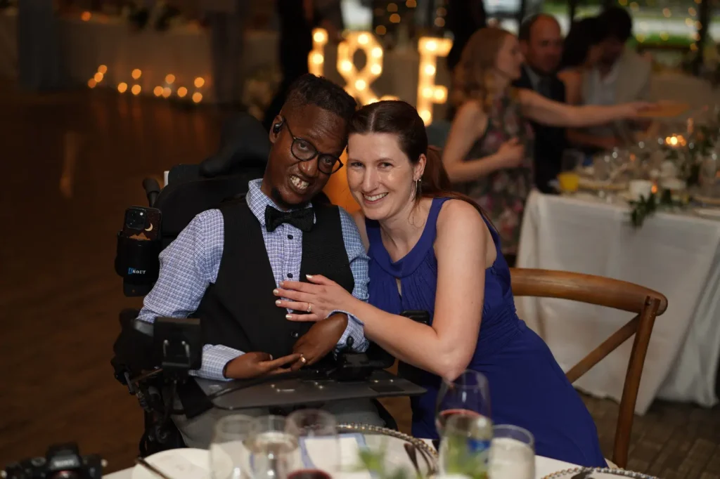 A man in a motorized wheelchair wearing a blue patterned shirt, black vest, and bowtie smiles alongside a woman in a vibrant blue dress at a warmly lit indoor evening event with decorative lights in the background.