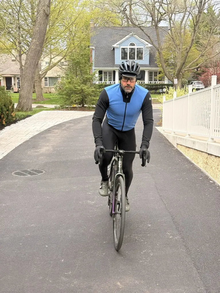 A cyclist wearing a black helmet, blue cycling vest and black athletic gear rides a road bike down a paved residential street. Trees, houses and a white fence line the quiet neighbourhood in the background.