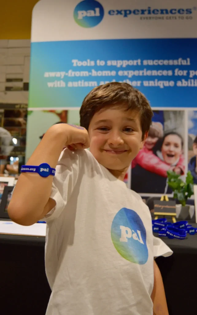 Charlie flexes his arm to show off a blue Pal Experiences wristband while standing in front of a banner for "Tools to support successful away-from-home experiences" at the Children's Museum of Phoenix.