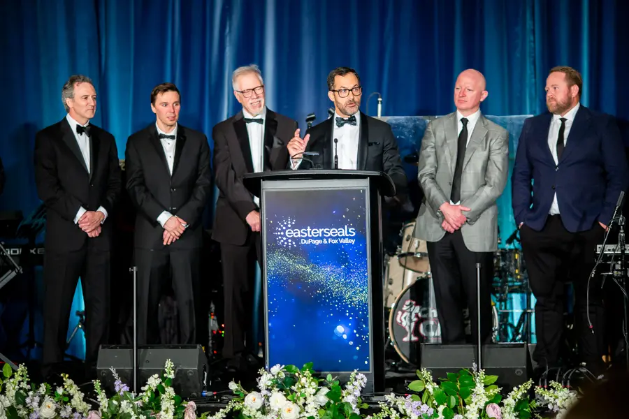 A man in a tuxedo speaking at a podium emblazoned with the "Easter Seals DuPage & Fox Valley" logo. Five other men in suits and formal wear stand behind him on a stage decorated with white and purple flowers.
