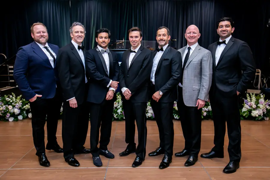 Seven men in various formal suits and tuxedos posing in a line for a group photo at the Easter Seals Gala. They are standing on a dance floor in front of a stage with a dark blue curtain.