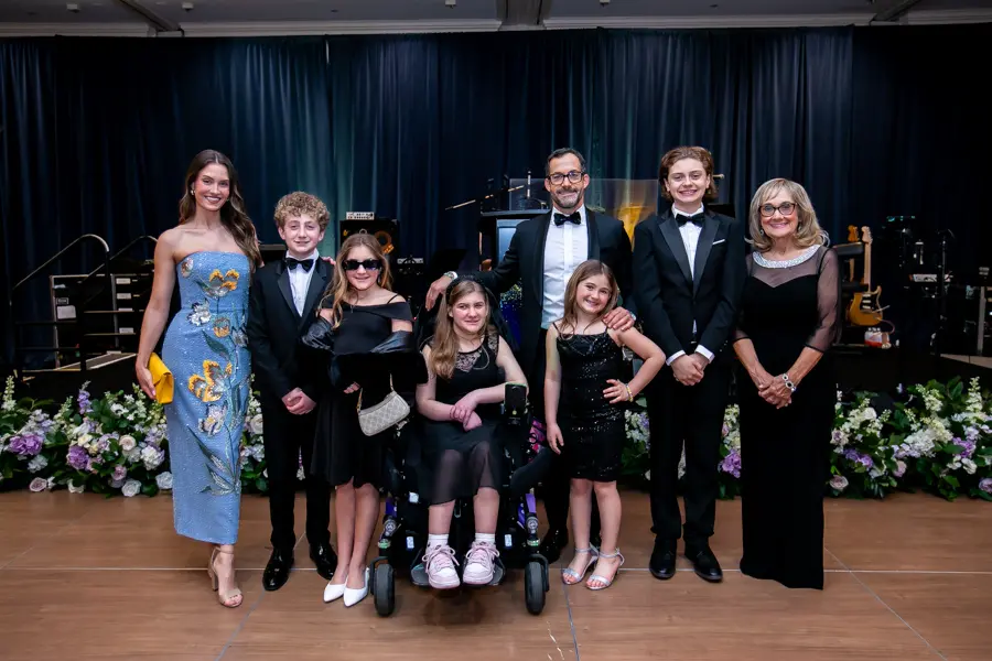 A multi-generational family posing together at the Easter Seals Gala. The group includes adults in formal wear, children in black-tie attire, and a young girl in a motorized wheelchair, all smiling in front of a blue draped backdrop and floral arrangements.