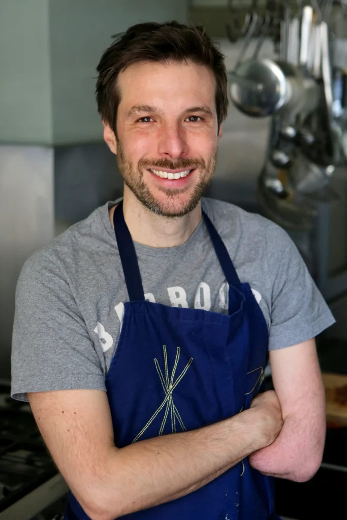 Portrait of a smiling man standing in a professional kitchen, wearing a blue apron over a grey T-shirt. He has short brown hair and light facial hair, with his arms crossed casually. Stainless steel kitchen utensils and cookware hang in the background.