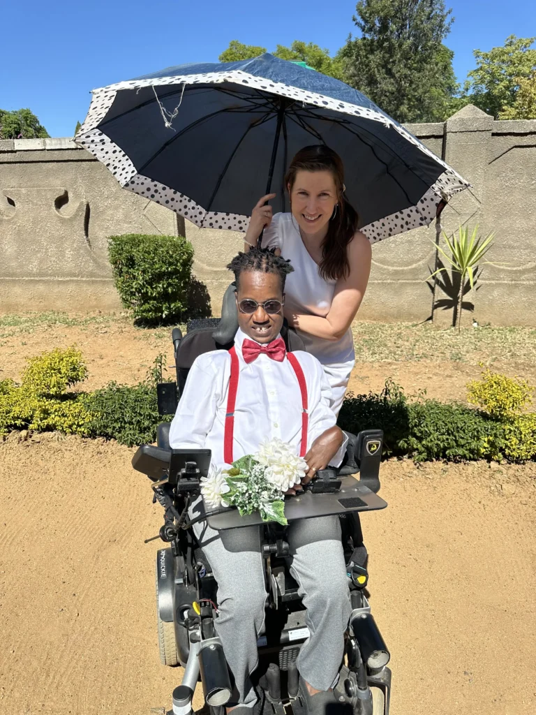 A smiling man in a motorized wheelchair wearing a white dress shirt, red bow tie, and red suspenders, while a woman in a white dress stands behind him holding a black and white polka-dot umbrella for shade in a sunny outdoor setting.