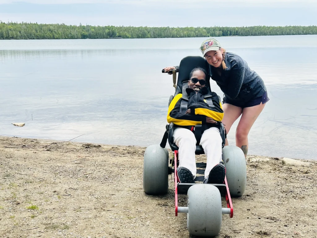 A man using an all-terrain beach wheelchair with large balloon tires sits on a sandy shore next to a calm lake, accompanied by a woman leaning in close and smiling for a photo against a backdrop of distant green forest.