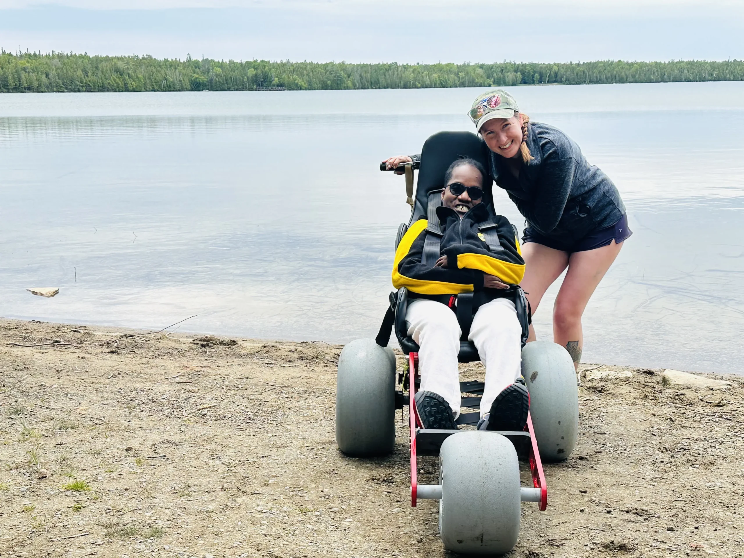 A man using an all-terrain beach wheelchair with large balloon tires sits on a sandy shore next to a calm lake, accompanied by a woman leaning in close and smiling for a photo against a backdrop of distant green forest.