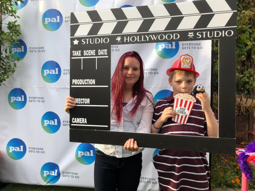 Jace and Rylan pose behind a large movie clapperboard prop at a Pal Experiences event, featuring "Studio Hollywood" branding and a "Pal: Everyone Gets to Go" backdrop.