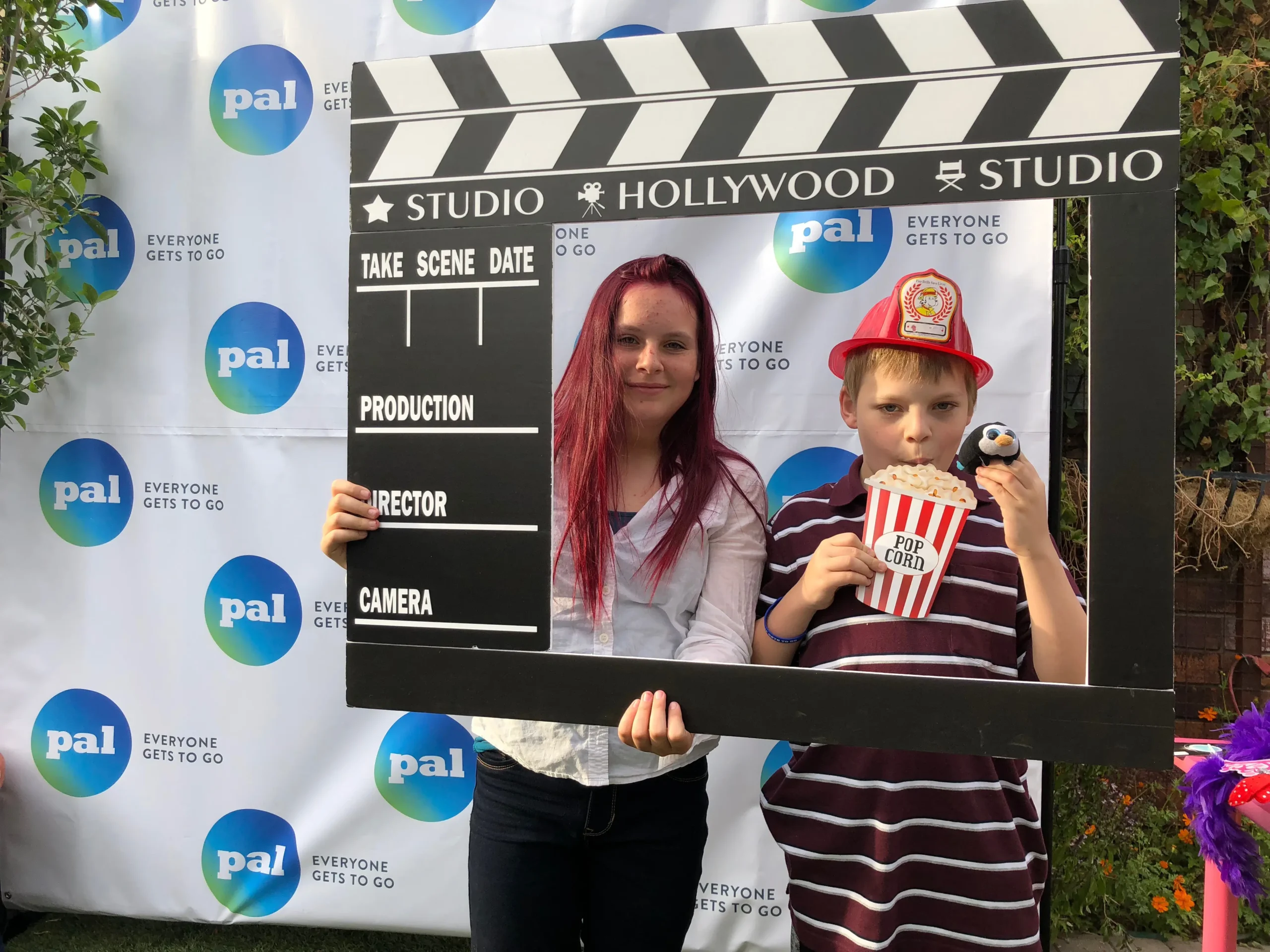 Jace and Rylan pose behind a large movie clapperboard prop at a Pal Experiences event, featuring "Studio Hollywood" branding and a "Pal: Everyone Gets to Go" backdrop.