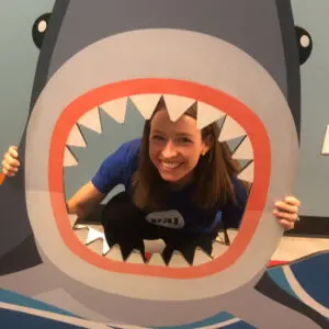 Melanie peeks through the open mouth of a large shark-shaped photo cutout, smiling at the camera during a Pal Experiences visit to an aquarium.