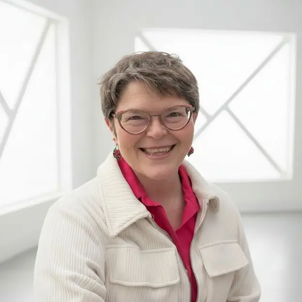 Headshot of a smiling woman with short gray hair and glasses, wearing a white corduroy jacket over a pink shirt, set against a bright, minimalist white background.