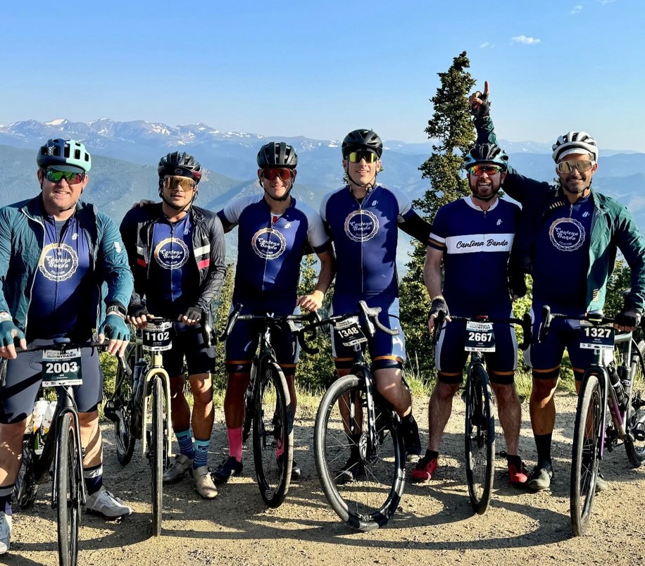 Six cyclists wearing matching blue cycling jerseys and helmets pose with their bikes on a mountain overlook during a race or endurance ride. Snow-capped mountains and trees stretch across the background under a clear blue sky. Race numbers are attached to the front of the bikes and the group appears to be taking a celebratory photo at a scenic viewpoint.
