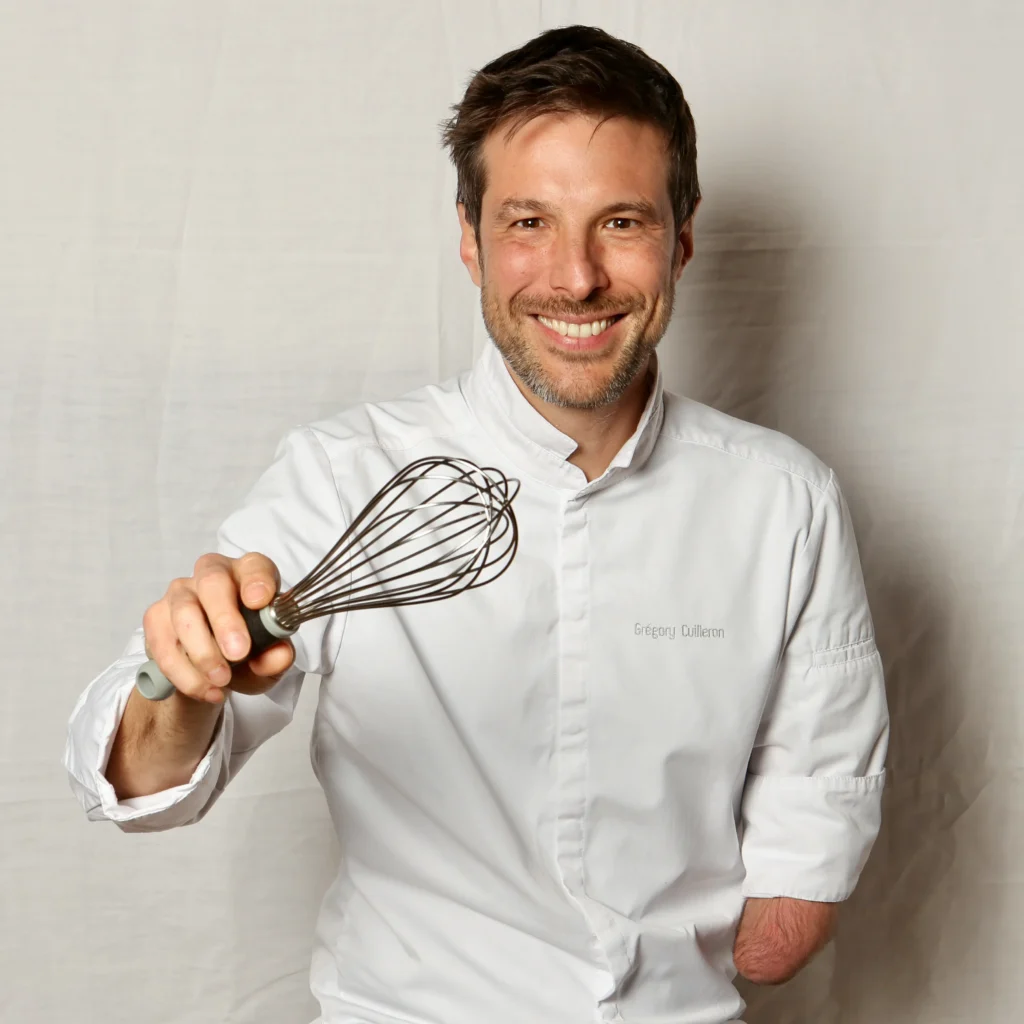 Professional portrait of French chef Grégory Cuilleron in a white chef's coat holding a wire whisk and smiling against a white background.