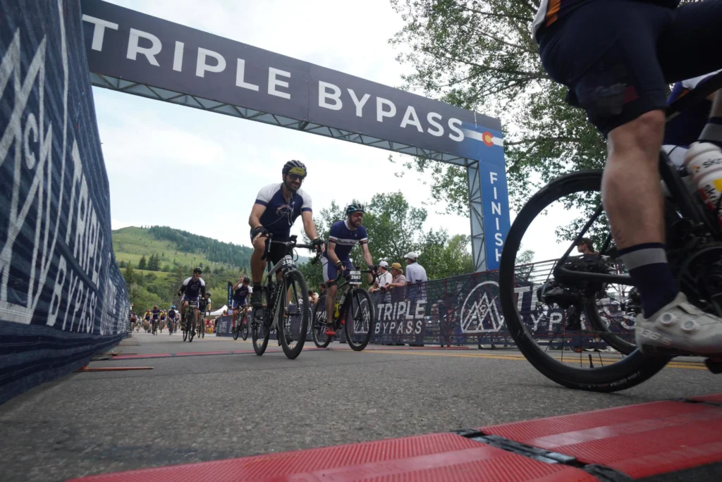 Cyclists crossing the finish line of the "Triple Bypass" ride under a large blue and white banner. The shot is taken from a low angle on the asphalt, showing the riders in motion.