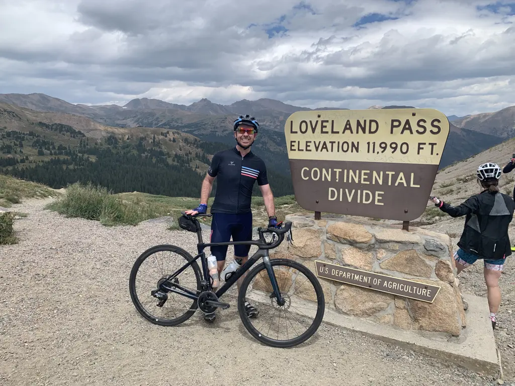 A male cyclist in a black and striped jersey smiling while leaning against his road bike next to a wooden sign marking "Loveland Pass, Elevation 11,990 FT, Continental Divide."