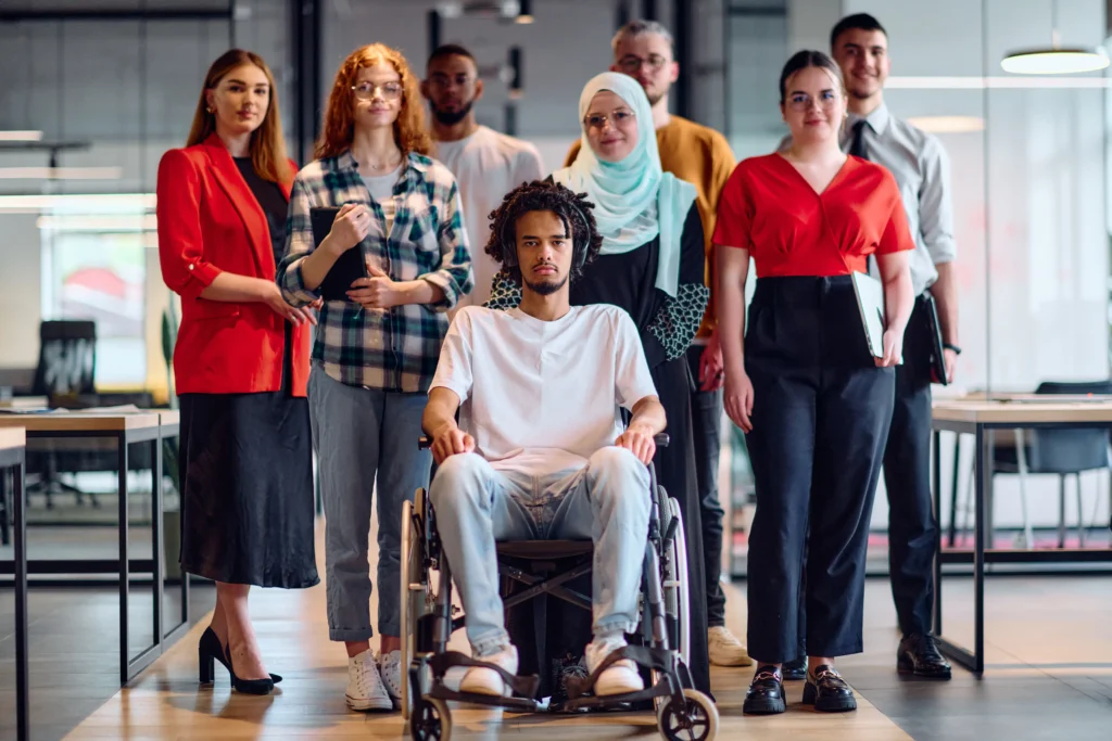 A diverse group of young professionals standing and sitting together in a modern, open-plan office. A man in a white t-shirt and wheelchair is positioned at the center foreground, surrounded by colleagues of various backgrounds, including a woman in a red blazer and a woman wearing a light blue hijab.